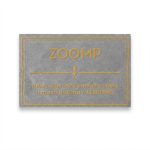 Etiqueta retangular de Zetex cinza com o logotipo da marca Zoomp aplicado em silk com alto relevo cinza e amarelo.